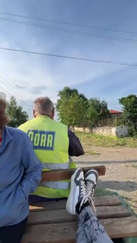 Four individuals are traveling in an open-air utility vehicle, possibly a rail cart, under a clear, sunny sky. An older woman with reddish-brown hair, wearing a blue zip-up jacket over a striped shirt, is seated on the left. To her right, an older man is seated with his back to the camera, wearing a bright yellow high-visibility vest with "DORR" printed in blue. Further back on the right, a young woman with long dark hair, wearing a light-colored sleeveless puffer vest and patterned grey leggings, is seated, at one point touching her hair. Her feet, clad in white and black sneakers, are elevated and resting on items in the cargo area, including a spare tire. The fourth individual's arm and leg are visible in the foreground, indicating they are either filming or a passenger. The vehicle moves through a semi-rural setting with houses, trees, power lines, and a dirt path or track visible in the background. A rhythmic metallic sound, consistent with a moving rail vehicle, is audible throughout the video.