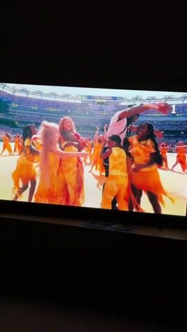 The video displays a television screen showing a live event. In the foreground, on the left, a woman with long blonde hair and a pink dress is holding a microphone and embracing a young child with blonde hair. They are surrounded by other children and adults, all wearing bright yellow and orange outfits. A tall individual with dark hair and a white t-shirt is being embraced by one of the children. In the background, a large stadium is filled with spectators. On the right side of the screen, a number "1" is visible. The video then cuts to a different scene where a man in a green jacket is speaking into a microphone on a stage, with a stadium full of people in the background. Text on the screen reads "#FIFACup".