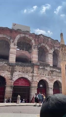 The video shows a Roman amphitheater on a sunny day. The structure is made of red and white stone with arches. There are several people walking around. One person in a red shirt is holding a phone and taking a picture. Other people are walking and looking at the amphitheater. There is a tall stone column in the foreground, with statues at the top. The sky is blue with some clouds. There are trees and buildings in the background.
