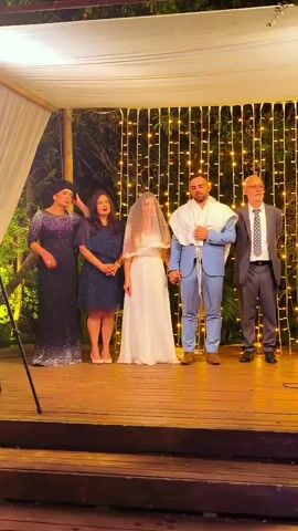 A group of five people stand on a wooden stage beneath a white canopy decorated with string lights. From left to right, a woman in a dark, sparkly dress and headscarf, a woman in a dark blue dress and headscarf, a bride in a white wedding dress and veil, a groom in a light blue suit holding a tallit over his shoulders, and a man in a grey suit and tie. The bride and groom are holding hands. The two women on the left appear to be placing their hands on the heads of the bride and groom respectively. The background is a wall of vertical strings of warm white lights. The overall scene appears to be a wedding ceremony.