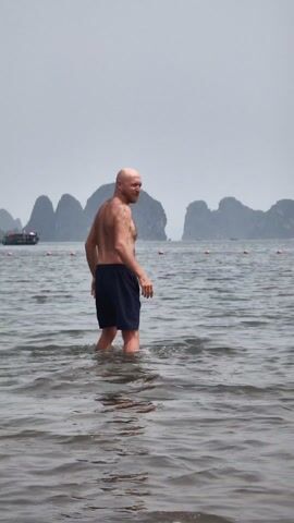 A medium shot shows a bald, shirtless man wearing dark shorts standing in the water, looking to the right. He turns around, showing his bare back to the camera. The water is shallow and choppy. In the background, there are several karst hills and a small boat with tourists, separated by orange buoys. The sky is overcast and gray.