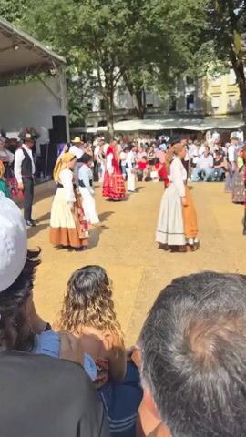 This is a video of a group of people in traditional attire dancing outdoors. The dancers are dressed in colorful skirts and blouses, some with vests and hats. They appear to be performing a folk dance in a square or park, with a stage and audience visible in the background. The dancers are moving in a circular formation, and some are holding hands. The environment is sunny, with trees and buildings in the background. The audience is seated and standing, watching the performance. The overall atmosphere appears festive.