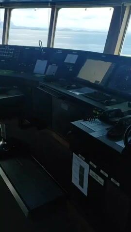 The image displays the interior of a ship's bridge, offering a view through the large front windows over the sea and a distant coastline or islands under a partly cloudy sky. The setting is daytime, with bright natural light illuminating the exterior scene while the bridge interior is relatively dimmer, enhancing the visibility of the control panels and screens. This suggests the vessel is operating in coastal waters near Kunak, Malaysia.

The main subject is the ship's control console, which is equipped with various instruments and displays. Several large digital screens are active, showcasing navigational information, likely including radar displays and electronic charts. On the right side of the console, a prominent red and green illuminated throttle control is visible, clearly set to "FULL AHEAD." Other marked positions on the throttle include "STOP," "SLOW," "HALF," "MAX" for ahead, and "ASTERN" with "SLOW," "HALF," "FULL" options. To the left of the throttle, a small steering wheel is integrated into the console.

Other visible objects on the bridge include communication devices, such as telephones, and numerous buttons, switches, and indicator lights across the panels, typical of a modern vessel's command center. A small digital display shows "12°" and "0.0," which could represent heading, speed, or other navigational parameters. A printed document is attached to the console, though its text is illegible. No people are visible in the frame. The active displays and the "FULL AHEAD" throttle setting indicate the ship is currently underway or actively preparing for movement at a significant speed.