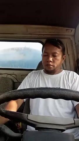 A man sits in the driver's seat of a truck, holding the steering wheel. He wears a white t-shirt. The interior of the truck is visible, including the steering wheel, dashboard, and seats. The man turns his head to look to the right.