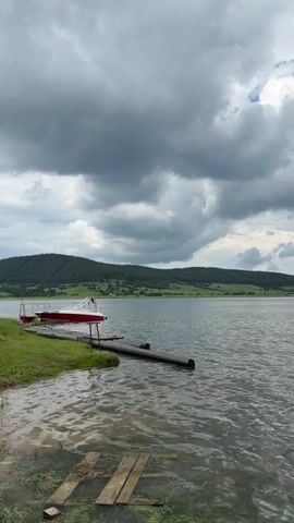 The video shows a body of water with a small boat resting on a wooden dock to the left. The water is still with subtle ripples, and the sky above is filled with a mixture of dark and bright clouds. In the background, green hills are visible under a partly cloudy sky, and a small island with trees can be seen in the distance on the right side of the frame. The camera pans slowly from left to right, revealing more of the lake and the distant landscape.