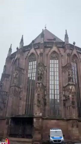 The video shows a large, ornate church-like building with tall windows and spires on a cloudy day. The building is made of a dark stone, possibly red or brown, and has intricate architectural details. A police van is parked in front of the building. A lamppost decorated with greenery is also visible in the foreground.