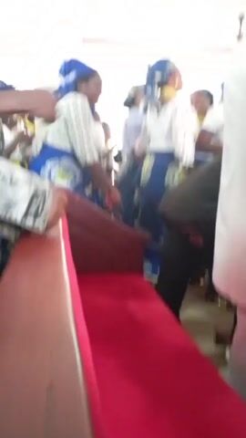 The video shows a group of people, primarily women, in what appears to be a church or religious gathering. Many of the women are wearing similar attire: white tops, blue skirts with a pattern, and matching blue head coverings. Some are clapping or moving to the music in the background. A woman in a red robe and a red head covering stands amongst the crowd, holding what seems to be a green book or pamphlets. A red velvet-covered pew is visible in the foreground, and the walls are painted white. Several men are also present, some wearing white robes, while one is dressed in black pants and a dark shirt. The general atmosphere suggests a celebratory or devotional event.