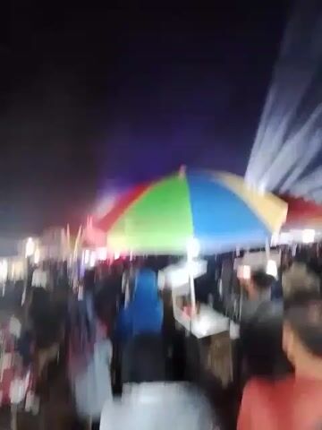 The video shows a crowded outdoor night market scene. A large, colorful umbrella is in the foreground, covering a food stall with a white cart. In the background, bright lights, possibly from a stage or light show, beam into the dark sky. Many people are visible, milling about and interacting. Some appear to be buying or selling goods at stalls. The overall atmosphere is lively and festive.