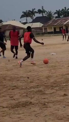 The media file shows a group of young men playing soccer on a dirt field. The ground is a light brown, sandy soil with some debris scattered on it. In the background, there are buildings with corrugated metal roofs and some palm trees, suggesting a tropical or subtropical location. The sky is overcast, indicating a cloudy day.

The main activity is a soccer game in progress. One player in a red jersey is dribbling the ball with his left foot. Another player in a dark blue jersey is approaching him, appearing to defend. In the distance, other players in red and white jerseys can be seen on the field. The players are dressed in athletic wear, including shorts and jerseys.

The time of day appears to be daytime, given the illumination. The weather seems to be mild and cloudy, without direct sunlight. The setting appears to be a rural or semi-rural area in Masaka, Nigeria, with simple structures and an unpaved playing surface common in such regions. The overall mood seems to be one of casual recreation and active engagement. There is no visible text in the media file.