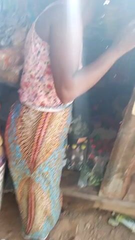 A woman wearing a floral print top and a long, patterned skirt stands in front of a small, wooden stall. The stall contains various items, including bottles, packages, and loose vegetables. The ground is dirt, and the structure is made of rough wood. The lighting is somewhat dim inside the stall.