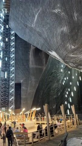 The video shows an interior view of a large cavernous space, likely a salt mine, with raw, rough-hewn salt walls. A massive, multi-story metal elevator shaft with illuminated panels runs vertically along one wall, reaching high into the darkness. Numerous elongated, bright blue lights are suspended from the ceiling at various heights, illuminating the cavern. In the foreground, a circular, elevated platform with a wooden railing and illuminated light fixtures contains what appears to be a food or souvenir stand. Several people are visible on and around this platform, including adults and children, some walking and others seated. More people are seen walking in the open space below the platform. The environment is dimly lit, with artificial lights providing the primary illumination.