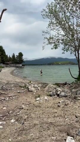 A young woman in a black bikini stands in shallow, greenish water near a rocky shore. Behind her, a large, cloudy mountain range rises above a distant treeline and buildings. The sky is overcast. The woman is facing away from the camera and appears to be looking out at the water.