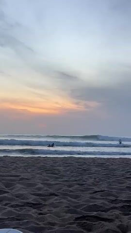The video shows a wide shot of a beach at sunset. The sky is filled with soft, pastel colors, ranging from pale blue at the top to a peachy orange near the horizon. There are some wispy clouds in the sky. The ocean is calm with gentle waves rolling onto the shore. In the distance, two surfers are visible in the water, one appears to be getting up on their board. The foreground of the video is dominated by dark sand, showing textured ripples. The overall mood is peaceful and serene.