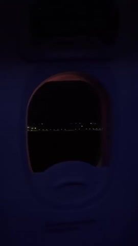 From inside an aircraft, the view through a small oval window shows a dark night scene. A faint line of lights stretches across the horizon, possibly indicating a distant city or coastline. The interior of the plane is dimly lit with a blue hue, highlighting the window frame and the lower part of the window. The scene is static, with no observable movement or interaction.