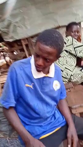 A man wearing a blue shirt with a white collar and a Puma logo sits on what appears to be a low platform of bricks. He looks down and then up at the camera. He is surrounded by wooden planks, cardboard boxes, and plastic sheeting, suggesting an area under construction or a makeshift shelter. Another person is partially visible in the background.
