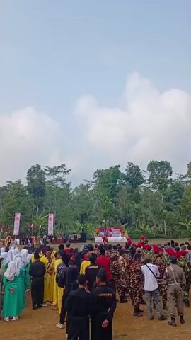 The video shows an outdoor gathering with a crowd of people in the foreground. The crowd consists of individuals in various attire, including what appears to be traditional clothing and uniforms. Some people are wearing hijabs, others are in yellow martial arts uniforms, and some are in camouflage and military-style uniforms. In the background, there is a stage with a red banner and several speakers, suggesting an event or ceremony. Behind the stage, a dense line of trees forms a natural backdrop. The sky above is partly cloudy. The ground appears to be dirt or sand. The overall impression is of a community event taking place in a somewhat rural or natural setting.