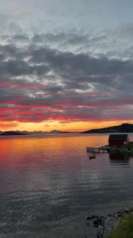 A serene video captures a breathtaking sunset over a calm body of water. The sky is ablaze with vibrant colors, ranging from fiery reds and oranges near the horizon to softer pinks and purples higher up. Dark, textured clouds are scattered across the sky, their undersides illuminated by the setting sun. The water reflects the sky's dramatic hues, creating a mirror-like surface. In the middle ground, a small red boathouse with a white-trimmed roof sits on a wooden pier extending into the water. A small dark boat is tied to the pier. In the distance, a silhouetted landmass with rolling hills can be seen against the colorful sky. The foreground shows the edge of the water with some rocks and pebbles visible along the shoreline. The overall atmosphere is peaceful and tranquil.