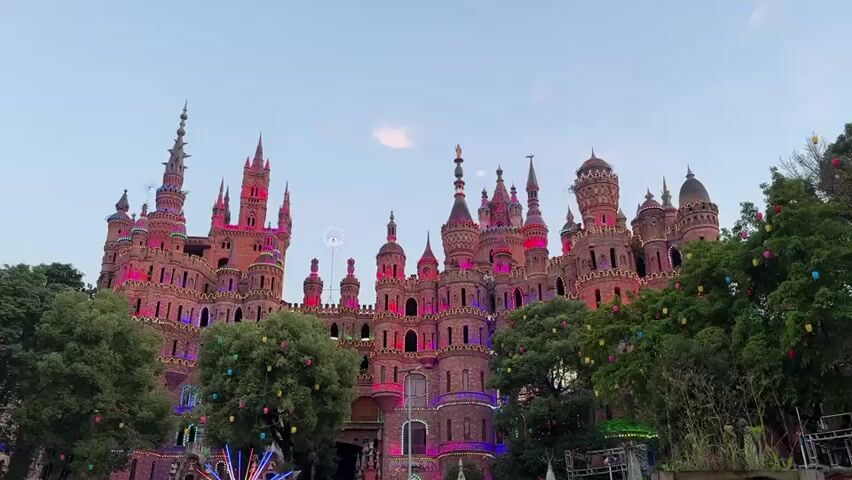 The video shows a large, ornate castle structure illuminated with red and pink lights, with several towers and spires visible against a light blue sky. Trees with colored lights hang between the castle and the viewer, partially obscuring the base of the structure. The scene suggests a festive or celebratory atmosphere.