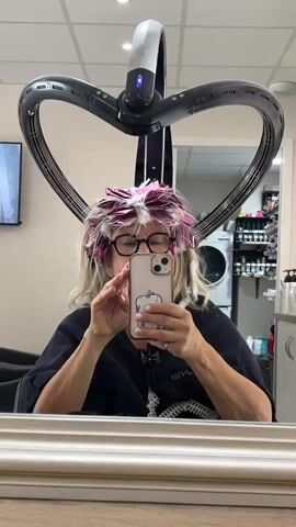 A person wearing glasses and a black smock is sitting in a salon chair with their hair covered in purple and white foils. They are holding a smartphone in front of them, taking a selfie. A large, heart-shaped hair dryer is positioned above their head. In the background, shelves stocked with hair products are visible, along with a washing machine. The scene appears to be a hair salon.