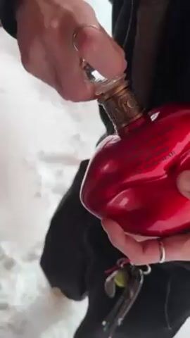 A person is opening a heart-shaped red bottle with a clear stopper outdoors on a snowy surface. The bottle has gold lettering on it. The person is wearing a black jacket and has rings on their fingers. Keys are hanging from their pants. They remove the stopper from the bottle.