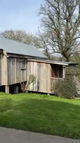 The camera begins pointed at a long, rectangular, single-story wooden building with a gray metal roof, situated on a grassy lawn. The building is elevated on posts, with some vegetation growing underneath. A small window with white curtains is visible. The camera then pans to the right, revealing a sidewalk and a person riding a bicycle on a path in the distance. The scene includes trees without leaves, a grassy area, and more wooden buildings in the background. The sky is light blue with some clouds.