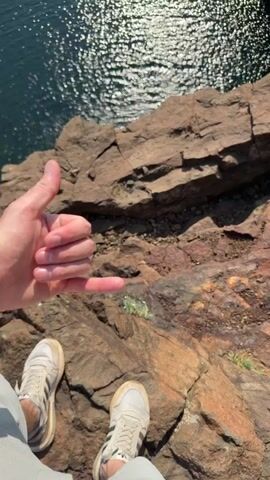 The video begins with a close-up view of a person's feet wearing white sneakers with black stripes standing on a rocky cliff edge. The person's hand is visible in the upper left corner, making a "shaka" or "hang loose" gesture. The camera then pans down and away from the person, revealing a vast body of dark blue water below, shimmering in the sunlight. The water appears to be a lake or a very calm section of a river, surrounded by steep, rocky cliffs. The perspective is from a high vantage point, looking down into the water and the surrounding natural landscape. The camera continues to pan, showing more of the cliff face, the water, and the shoreline. Towards the end of the video, a small waterfall can be seen cascading down one of the rocky inclines into the water. The scene is predominantly natural, with no man-made structures visible other than the implied presence of a path or viewing area at the cliff's edge where the person is standing. The overall impression is one of a scenic overlook of a natural body of water and a rugged, rocky environment.