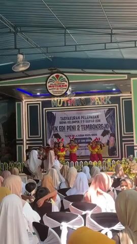 A group of children in colorful costumes are performing a dance on a stage in front of an audience. The stage is decorated with banners and lights, and there is a large sign that says "MUTIARA PARK". The audience is seated in chairs and many of them are wearing headscarves. The event appears to be a graduation or year-end celebration for a kindergarten.