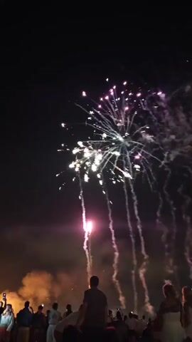 The video shows a fireworks display at night. A crowd of people are watching from below, silhouetted against the bright explosions in the sky. The fireworks are colorful, with bursts of red, white, and green light illuminating the dark sky. Smoke trails descend from the fireworks, adding to the dramatic effect. The scene is festive and celebratory, capturing the excitement of a fireworks show.