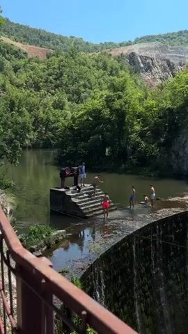 This video captures a group of young men enjoying a sunny day by a waterfall in Uzice, Serbia. The scene is set amidst lush green trees and rocky cliffs, with a dam forming a natural pool above a cascading waterfall. The water appears calm and inviting, reflecting the bright sky.

Several individuals are seen interacting with the water and the dam structure. On the dam itself, one person stands at the top, seemingly supervising or about to jump, while others are positioned on the stepped concrete structure leading down to the water's edge. Some are wading in the shallows, while others appear to be preparing to enter the water. One person is seated on the edge, looking out at the scene. The overall atmosphere is one of leisure and recreation, with the warm weather encouraging outdoor activities.

Notable details include the presence of a large, red industrial mechanism on the dam, possibly part of its original function. The rocky terrain and dense foliage suggest a natural, somewhat rugged environment. The sunlight indicates it is likely midday or early afternoon. The men are dressed casually in swimwear, suggesting a relaxed and informal gathering. There is no visible text in the video. The activity appears to be a casual swim and hangout at a picturesque natural spot.
