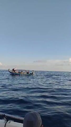 The video shows a clear blue sky and the ocean with moderate waves. A boat is in the middle distance, carrying several people. Another boat is to the left. The camera is positioned on another boat, looking out towards the ocean and a distant island. There are people on the boat in the foreground, some wearing headscarves. The overall impression is of a pleasant day at sea.