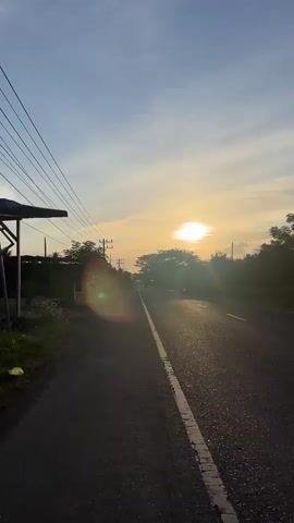 The video shows a road with a sunset in the background. The sun is partially obscured by trees. The road is paved and has a white line down the center. There are power lines on the left side of the road, and trees and vegetation on both sides. There is a slight lens flare in the foreground. The sky is blue with some clouds. The video appears to be filmed from a moving vehicle, as evidenced by the slight blur and the changing perspective. There are no people or animals visible in the video.