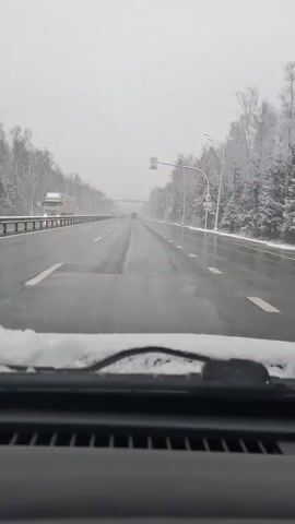 The video presents a first-person perspective from inside a car driving on a multi-lane highway during winter. Light snow is actively falling, and a thin layer of snow rests on the base of the vehicle's windshield and wipers.

The road surface is wet, with clear white dashed and solid lane markings. Snow has accumulated on the road shoulders and atop the guardrails that line both sides of the highway. Dense forests, with trees heavily laden with snow, flank the road.

A white truck with illuminated headlights is visible on the left side of the road, heading in the opposite direction. Several other distant vehicles can also be seen ahead, some with their headlights on, traveling in both directions. An overhead gantry structure spans the highway in the mid-distance.

Suspended from an overhead pole on the right side of the road, a circular white speed limit sign with a black border prominently displays the number "100" in black, indicating a 100 km/h speed limit. Streetlights are visible along the highway, though the overcast sky provides diffuse daylight. The scene depicts a typical winter driving experience in snowy conditions, consistent with the Yamuga, Russia context.