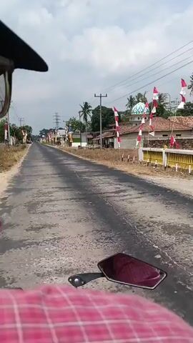 The video shows a first-person perspective from someone riding a motorcycle down a paved road. The rider is wearing a black hat, glasses, and a black face mask. The motorcycle has a pink and white checkered pattern on its seat and a red handlebar grip is visible. The road is lined with small buildings, trees, and utility poles. On the right side of the road, there is a building with a light blue dome, possibly a mosque. Numerous Indonesian flags are displayed along the roadside. The sky is cloudy, and the scene appears to be in a rural or suburban area.