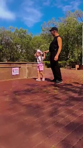 A man in a black sleeveless shirt, black pants, and a black baseball cap stands to the right of a young girl. The girl wears a white t-shirt, pink shorts, and a pink hat. They are in a park with a brick pathway and trees. The man holds the girl's left hand. The girl is looking down and to her left. The man is looking down and to his left. A sign with a lightning bolt symbol is visible on a stone wall to the left of the girl. The sky is blue with some clouds.