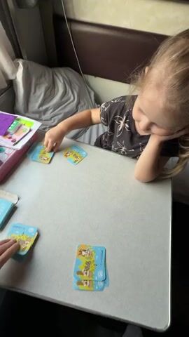 A young blonde-haired child and an adult's hand are playing a card game on a light-colored table inside a train carriage. The child, wearing a dark patterned shirt, is focused on the cards, resting her chin on her hand, and is in the process of placing a card. The adult's hand is also visible, placing a card down.

The game involves small square cards with various illustrations, such as anchors, sailboats, strawberries, rainbows, and shoes. Many of the cards feature the "АЭРОФЛОТ" (Aeroflot) and "SELECT" logos, suggesting they might be part of an airline-themed game or set. Several stacks of cards are visible on the table, and a purple box along with small blue boxes are also present, likely containing more game components.

The setting appears to be a train compartment, with a bed or seat covered in grey bedding visible in the background, typical of a sleeper car. The lighting suggests it is daytime. The child appears engaged in the game.