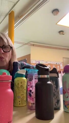 A woman with short blonde hair and glasses is standing behind a table filled with various colorful water bottles. She makes a kissing face, then sticks out her tongue while pointing to the bottles with both hands. The background appears to be a classroom or office space, with a yellow support beam, posters on the wall, and ceiling lights visible.