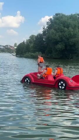 The video shows three people on a red pedal boat shaped like a car on a body of water. The person in the back of the boat is standing up and appears to be steering. The other two people are seated and wearing orange life vests. The pedal boat is moving through the water, leaving a wake behind it. The water is choppy, and there are trees and houses in the background. The sky is blue with some white clouds. The lighting suggests it is daytime.