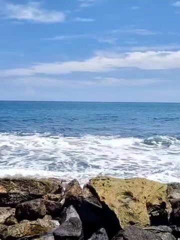 The video shows a rocky shoreline with waves crashing against the rocks. The ocean is blue and there are some clouds in the sky. In the distance, there is a town with buildings and a harbor. The camera pans from left to right, showing more of the coastline and the town.