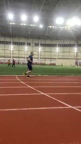 The video shows a man running on a red indoor track. He is wearing a dark blue shirt, dark shorts, and bright yellow shoes. There are white lines marking the lanes on the track. To the side of the track is a grassy area with a white soccer goal. Several people are standing and sitting in the background, near the grassy area. The man is running away from the camera. The building has a high ceiling with lights and a white grid structure.