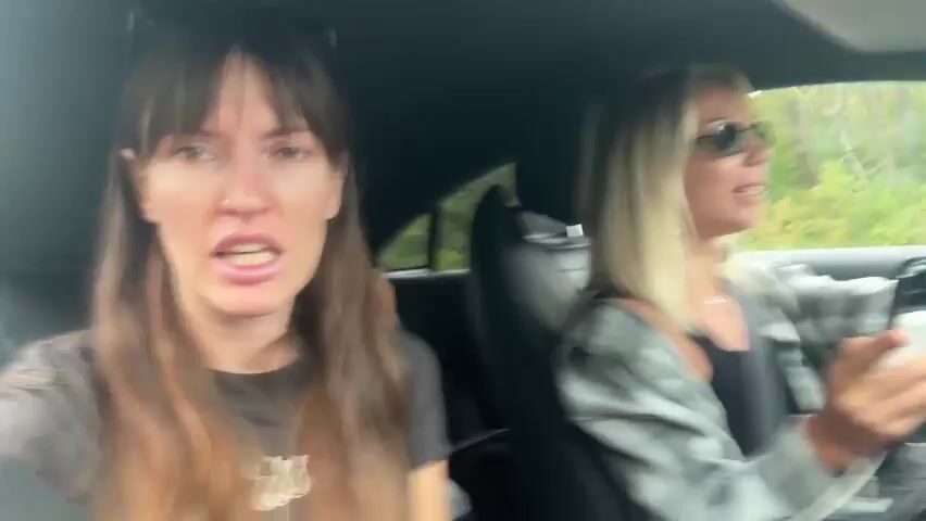 Two women are in a car. The woman in the foreground is facing the camera and speaking. She has long brown hair and is wearing a gray t-shirt. The woman in the background is driving. She has blonde hair and is wearing sunglasses and a plaid shirt. She is holding a white phone in her right hand. The car is moving, and trees and greenery are visible outside the windows.