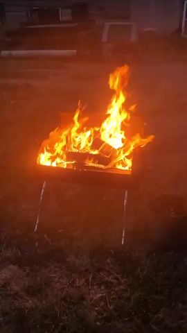 The video shows a campfire burning in a metal fire pit. The fire pit is black and has four legs. The fire is bright orange and yellow, with flames reaching upwards. There are logs burning inside the fire pit. The ground around the fire pit appears to be covered in grass and dirt. The background is dark and out of focus, suggesting it is nighttime or dusk. There is a dark object, possibly a bucket, to the right of the fire pit.