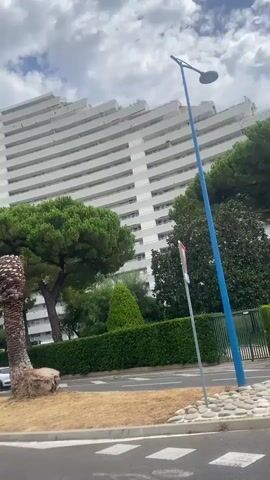 The video pans across a street with cars parked along the side, in front of a large, modern, white apartment building with many balconies. There are trees and greenery in the foreground and around the building. A few palm trees are visible, one of which appears to have been cut down to a stump. The sky is cloudy.
