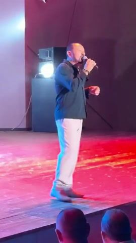 A man with short hair and a beard, wearing a dark shirt and light pants, stands on a stage with red and blue lighting, holding a microphone in his right hand. He gestures with his left hand as he sings. In the background, there are black sound equipment and a speaker.