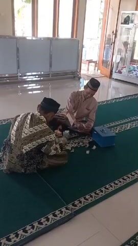 Two men are sitting on a green carpet in what appears to be a room with tiled floors and large windows. Both men are wearing traditional Muslim attire, including peci hats. One man on the left is wearing a black and gold patterned robe and is seen counting money. The man on the right is wearing a beige robe and is also counting money, smiling and looking at the other man. Between them are piles of paper money, a blue donation box, and what looks like a clear container with money inside. The room has white partitions in the background and a glass cabinet on the right. Sunlight streams in through the windows, creating shadows on the floor. The overall atmosphere appears to be one of calm and perhaps an act of charity or financial counting for a religious purpose.