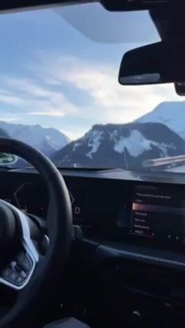 The video shows a view from inside a car, likely a BMW, with the dashboard and steering wheel visible. The car is in "D" drive mode. A digital display on the dashboard shows settings options. The driver's hand is visible on the steering wheel. The exterior view through the windshield reveals snow-covered mountains under a blue sky with scattered clouds.