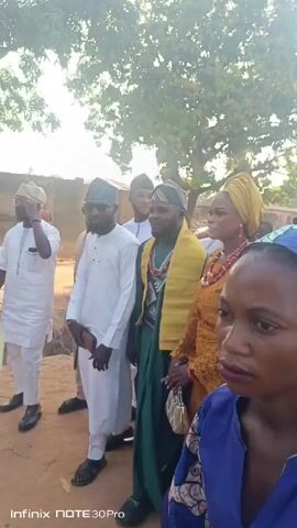 A video shows a group of people gathered outside. Most of the people are wearing traditional clothing. Two men are wearing white robes and matching hats, with one holding a pair of sandals. Another man in a green robe with a yellow shawl walks next to a woman in a golden yellow lace dress, wearing a yellow head wrap and red coral necklace. Another woman stands to her right wearing a blue dress with a patterned head wrap. A tree stands behind the group. The camera pans across the scene as people stand and talk. The setting is outdoors, with a dirt ground.