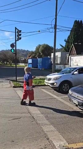 A clown in red pants and a blue and white shirt is juggling three green balls in a crosswalk in front of cars. The crosswalk has a green walk sign lit. The sky is blue with many power lines visible. There are also some trees, buildings and shipping containers visible in the background.