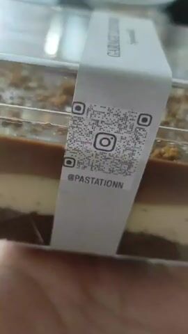 The video shows a close-up, hand-held shot of a dessert in a clear plastic container. The dessert is layered, with a dark base, a light middle layer, and a dark chocolate top layer. The top layer is sprinkled with crumbled cookie or cake pieces. A white paper band wraps around the container, displaying the text "GARAGE LOUNGE" and "Spoonfull." There is also a QR code and an Instagram logo on the band. The lighting is diffused, and the background is blurred.