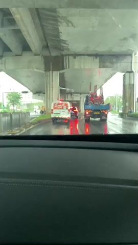 The video shows a view from inside a vehicle driving under an elevated highway or bridge structure. The road is wet, indicating it is raining. Several vehicles are visible on the road ahead. In the immediate foreground, the dashboard of the vehicle is visible. Further ahead, a white van with red markings on its rear is driving, followed by a smaller red vehicle. To the right of these vehicles is a large blue truck carrying a red and blue load, possibly construction equipment. Further down the road, other vehicles are visible, including what appears to be a yellow truck in the distance. Traffic lights are visible, with red lights illuminated for the direction the camera is facing, suggesting the vehicles are stopped or slowing down. The concrete structure of the elevated roadway is prominently visible overhead, with some water stains and drips on its underside. The overall environment appears to be urban or suburban.