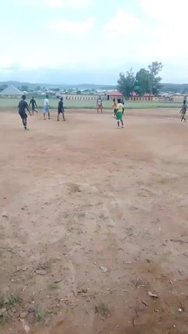 This is a video of a soccer game being played on a dirt field. There are several people on the field, some of whom appear to be playing soccer. The field is surrounded by trees and buildings in the background. The sky is overcast. The video is from a first-person perspective.